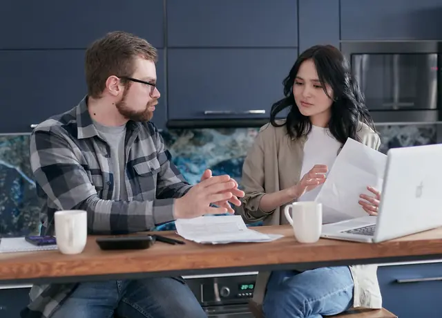 Two people at a kitchen table going through paperwork on a laptop